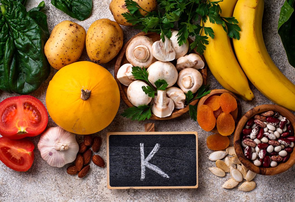 Verschiedene Lebensmittel wie Bananen, Orangen, Pilze und Nüsse rund um eine kleine Tafel mit der Aufschrift „K“