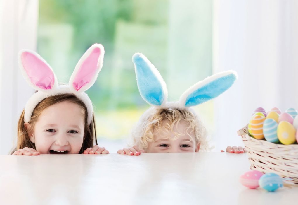 Zwei Kinder mit bunten Hasenohren schauen über einen Tisch mit bemalten Ostereiern