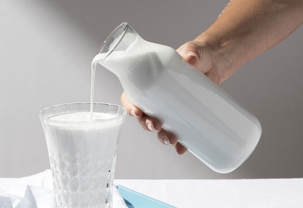Hand gießt frische Milch aus einem Glasgefäß in ein Glas.