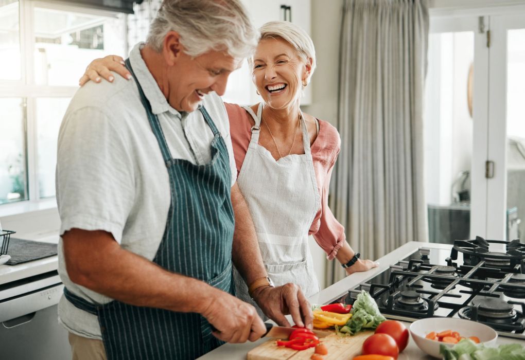 Älteres Paar steht in der Küche und bereitet lachend frisches Gemüse zu