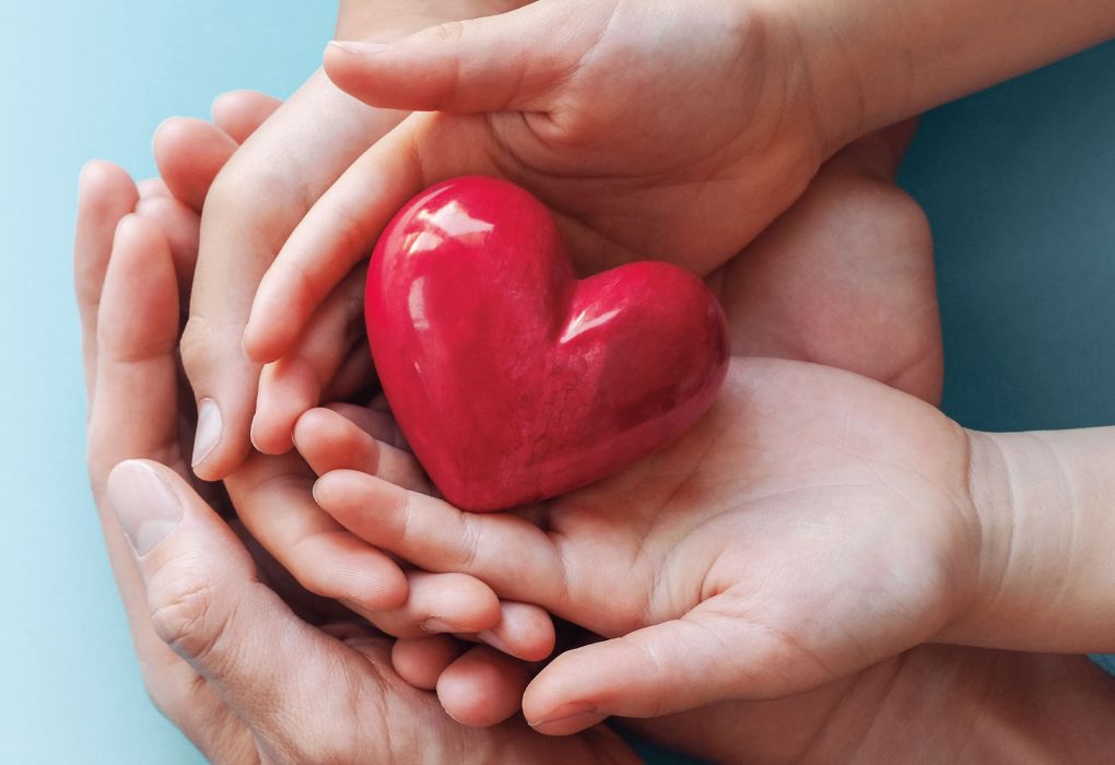 Rote Herzfigur wird von zwei erwachsenen Händen und einer Kinderhand behutsam gehalte