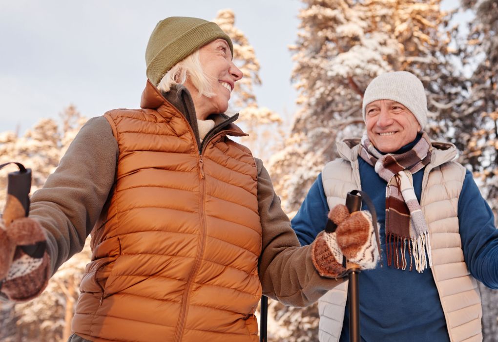 Älteres Paar spaziert lachend im verschneiten Wald und genießt Bewegung an der frischen Luft