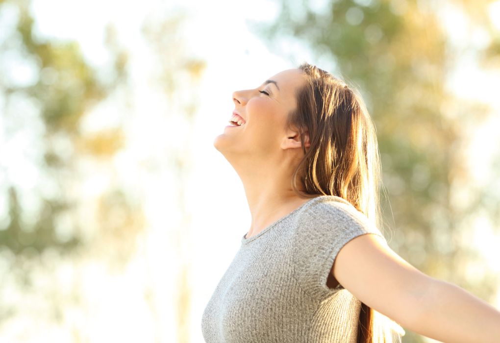 Junge Frau steht draußen, atmet tief ein und schließt die Augen, Symbol für Entspannung.