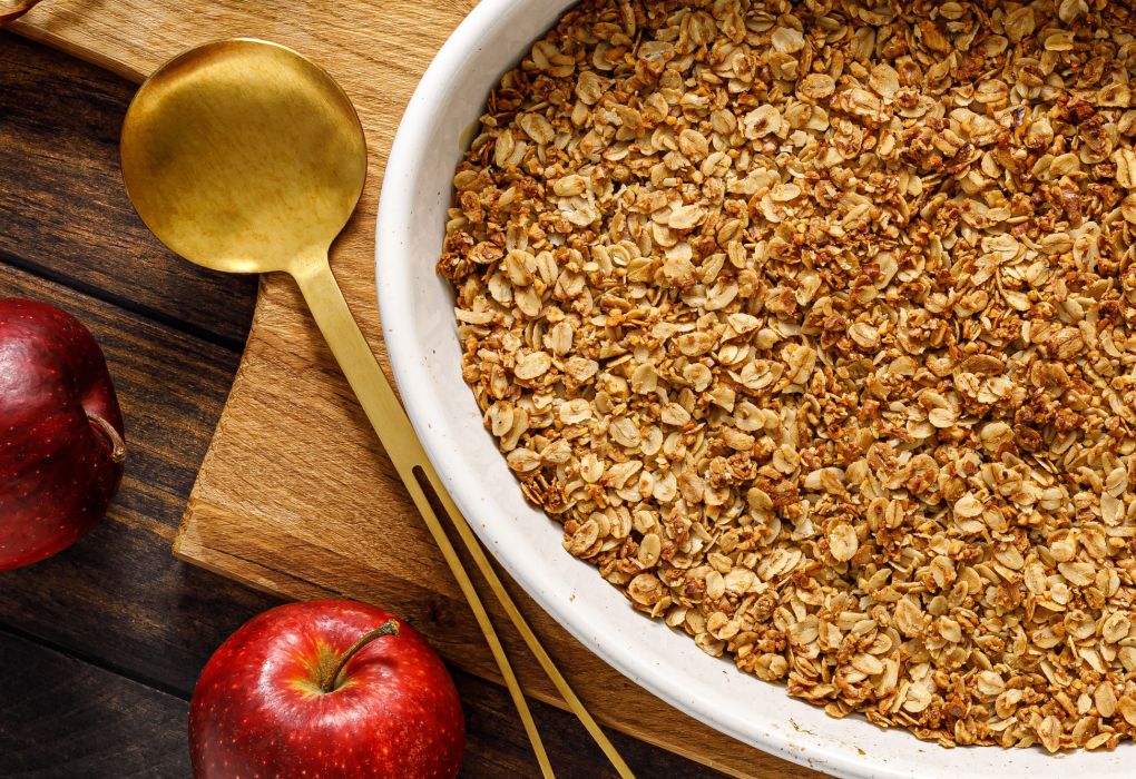 Auflaufform mit gebackenem Apple Crumble, daneben frische Äpfel und Zimtstangen