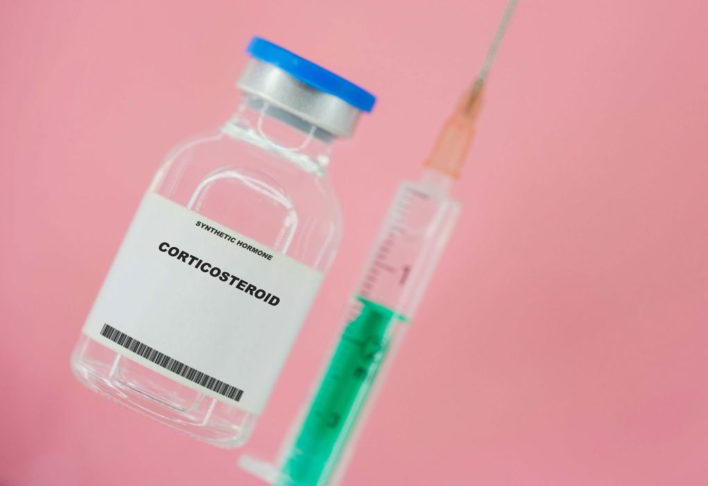 Medizinische Ampulle mit Aufschrift Corticosteroid neben aufgezogener Spritze vor rosa Hintergrund