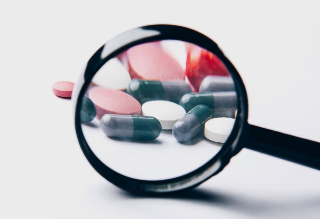 Mehrere Kapseln und Tabletten liegen unter einer Lupe auf weißem Hintergrund