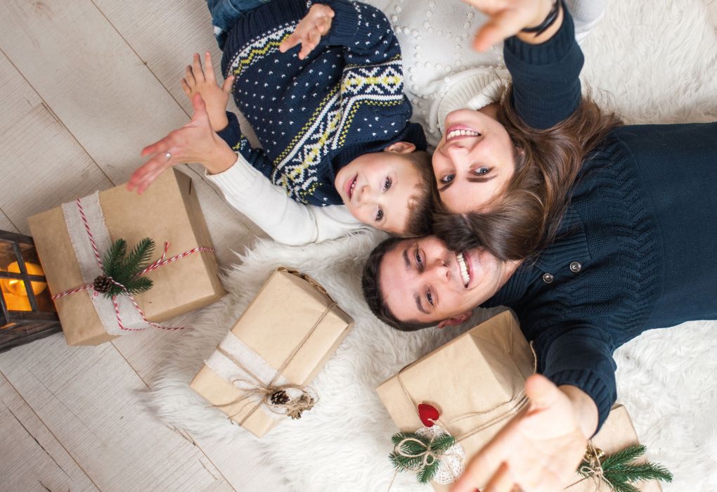 Eltern mit Kind liegen lachend auf dem Boden neben einem Weihnachtsbaum und Geschenken