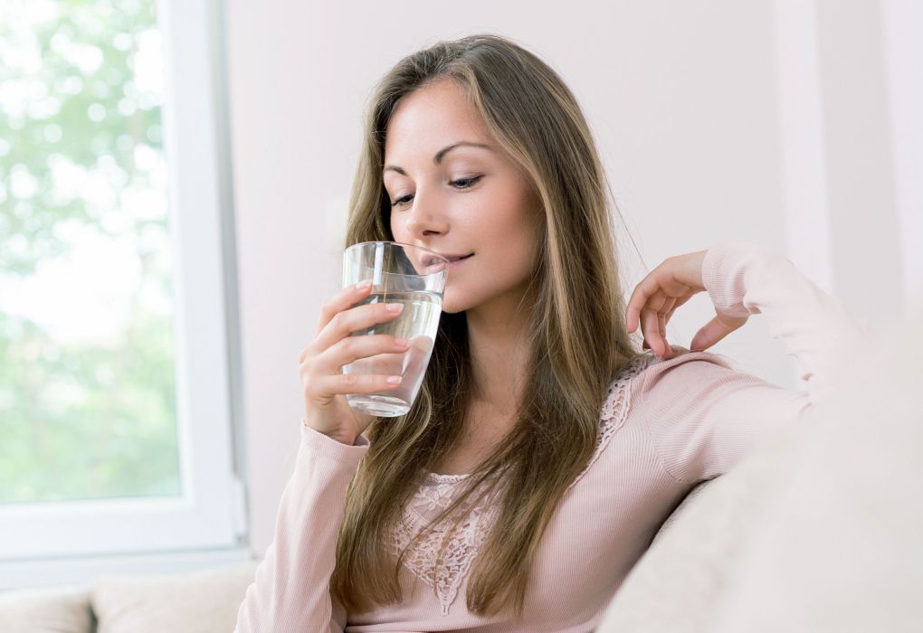 Junge Frau sitzt entspannt am Fenster und trinkt ein Glas Wasser