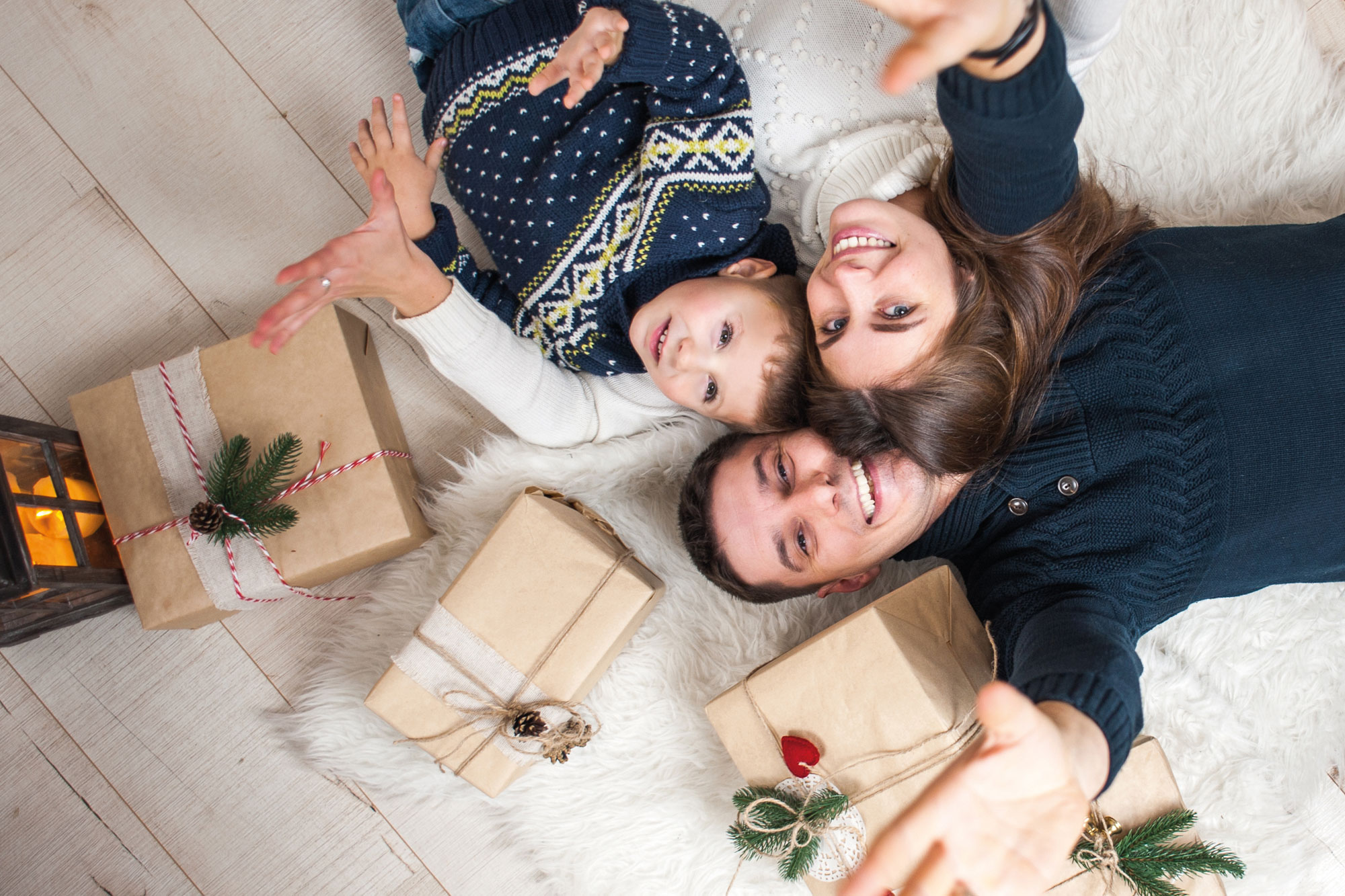 Eltern mit Kind liegen lachend auf dem Boden neben einem Weihnachtsbaum und Geschenken