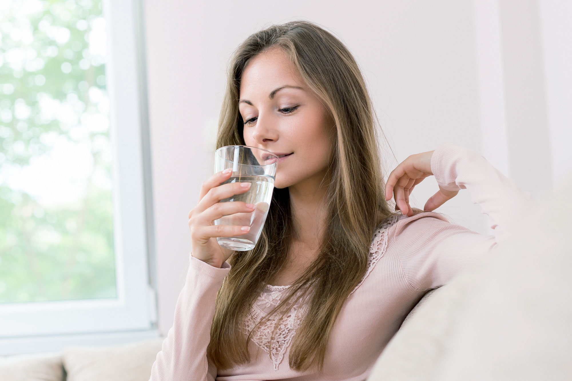 Junge Frau sitzt entspannt am Fenster und trinkt ein Glas Wasser