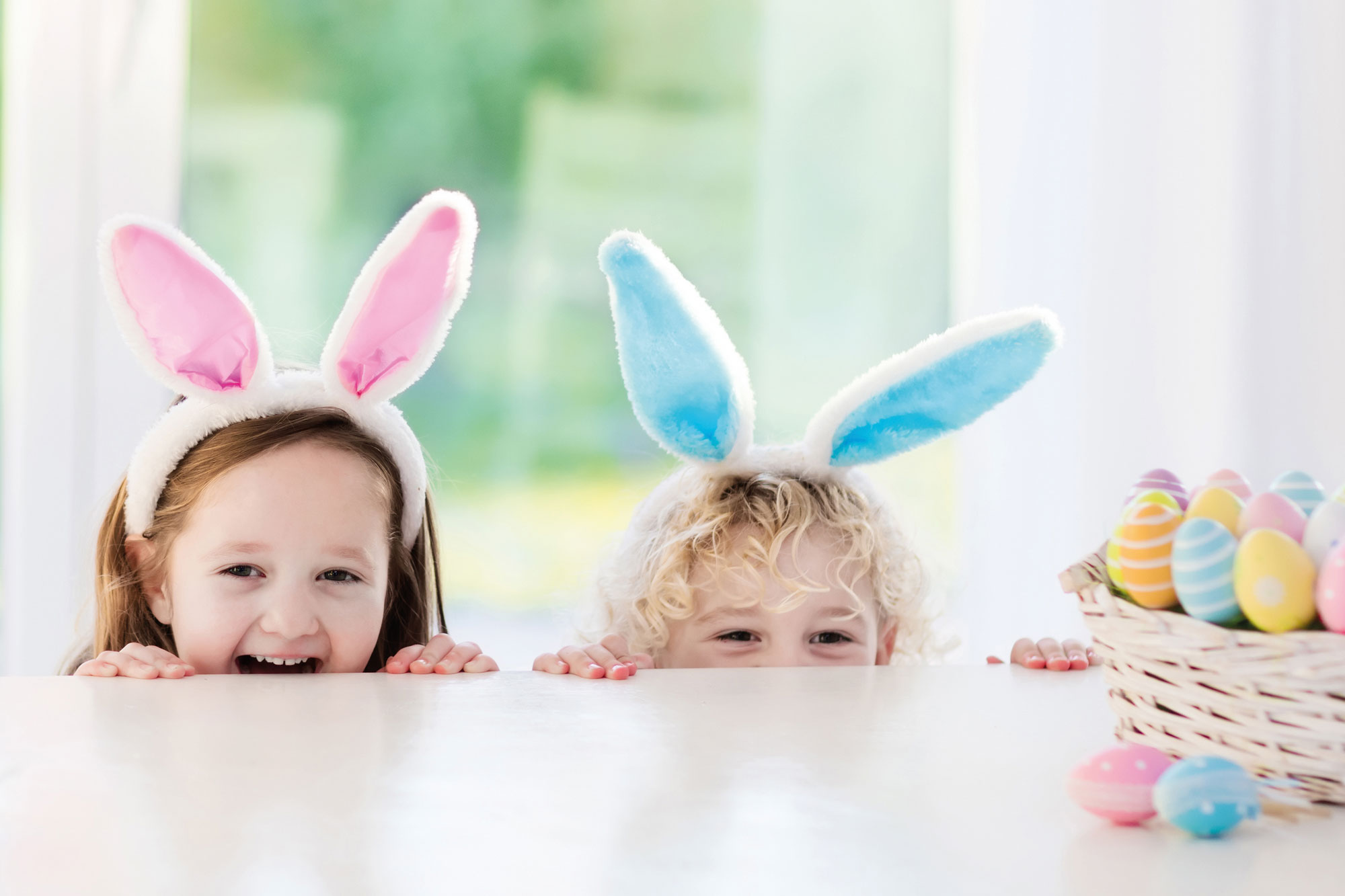 Zwei Kinder mit bunten Hasenohren schauen über einen Tisch mit bemalten Ostereiern