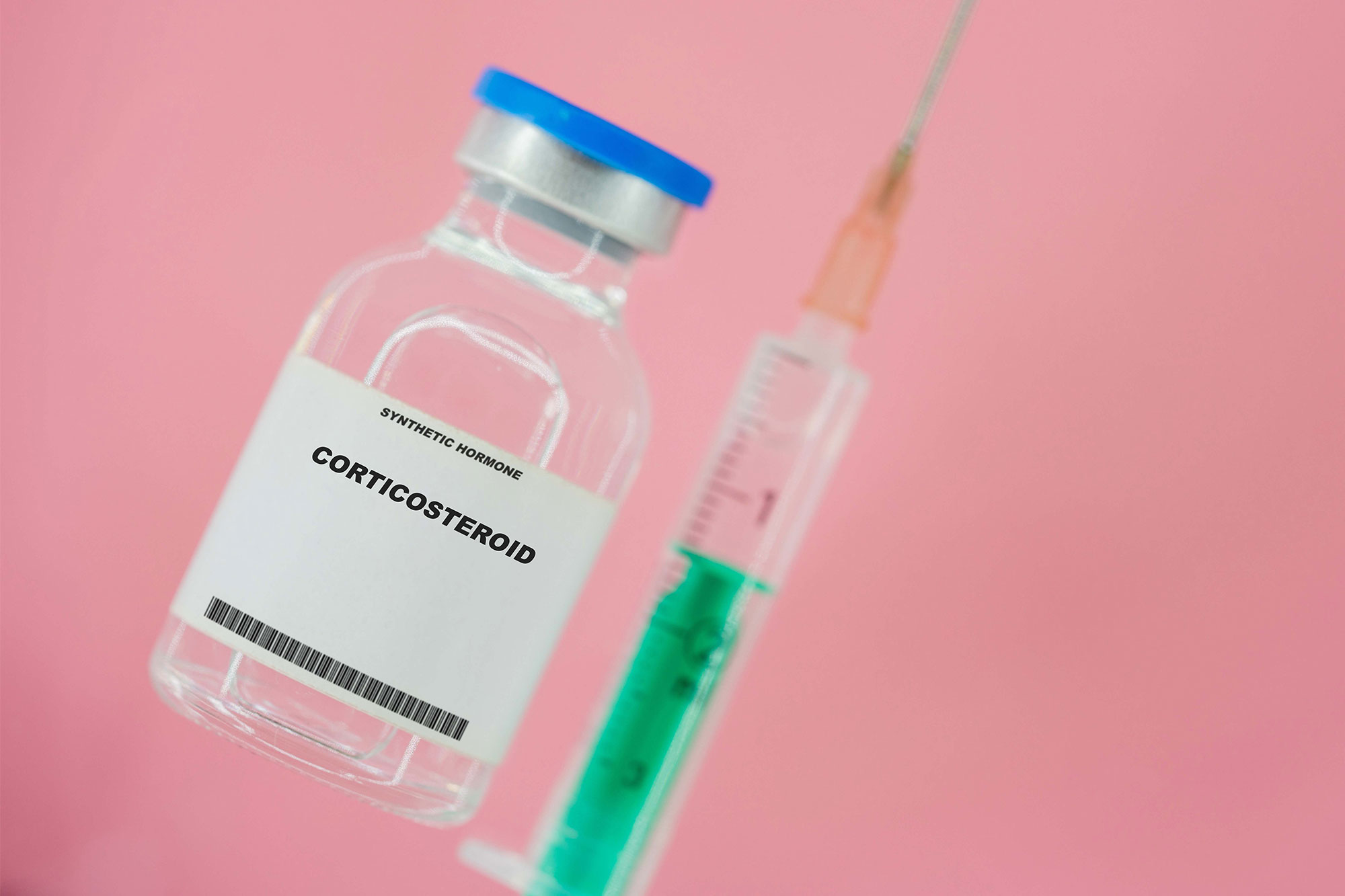 Medizinische Ampulle mit Aufschrift Corticosteroid neben aufgezogener Spritze vor rosa Hintergrund