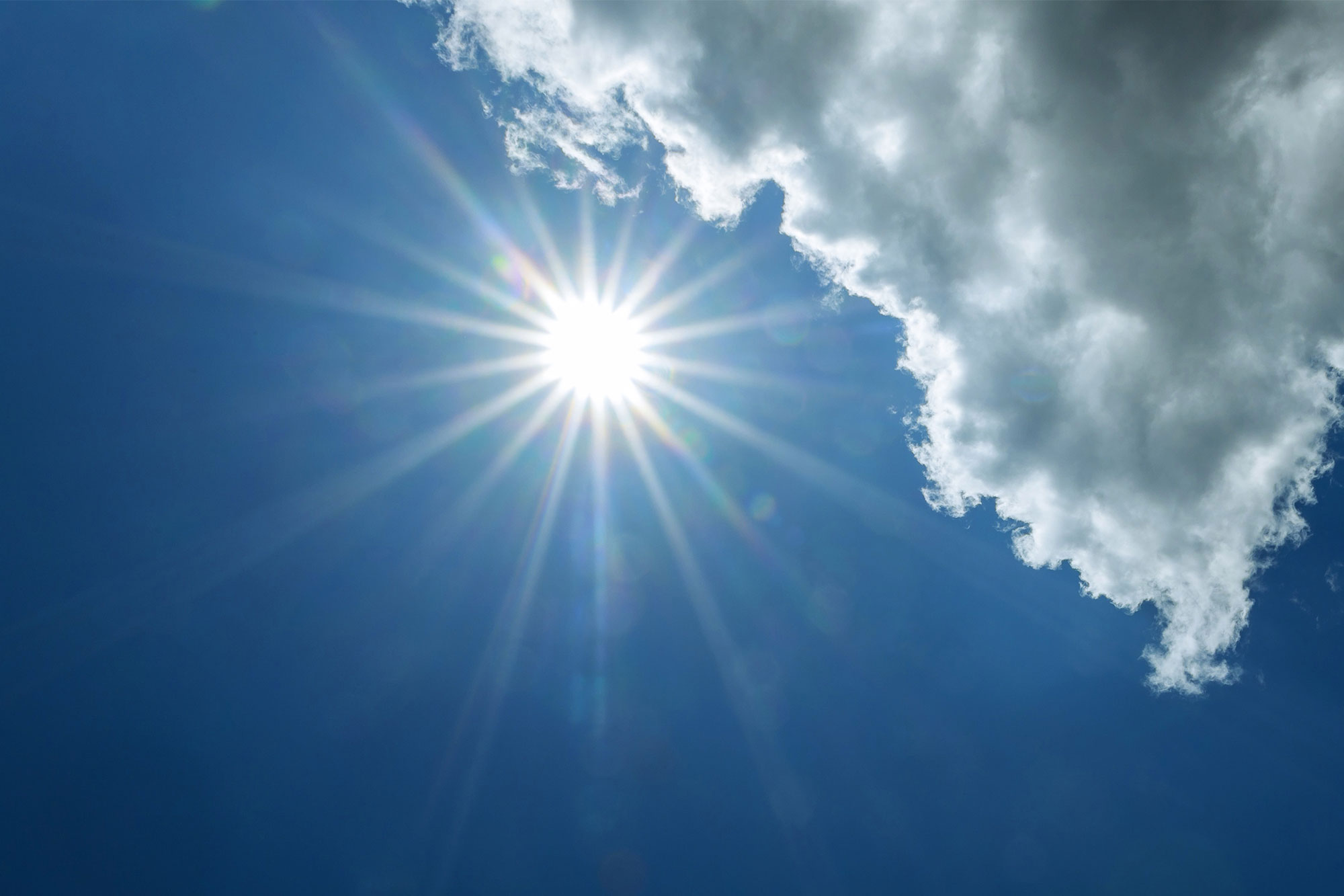 Starke Sonnenstrahlen brechen durch weiße Wolken am blauen Himmel