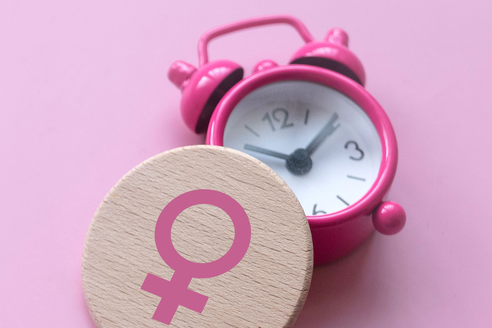 Holzscheibe mit Frauenzeichen neben rosa Wecker auf pinkem Hintergrund