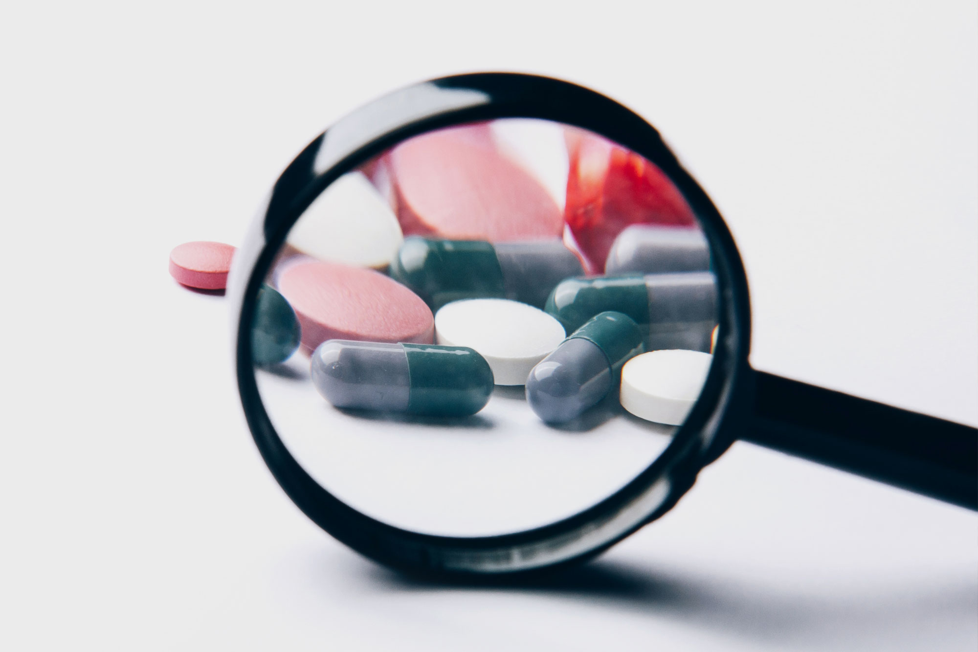 Mehrere Kapseln und Tabletten liegen unter einer Lupe auf weißem Hintergrund